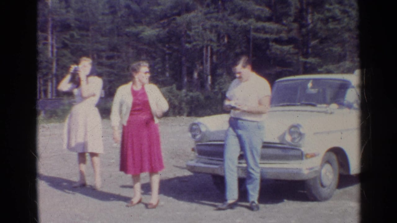 Two women and a man stand beside a white vintage sedan near a forest road.