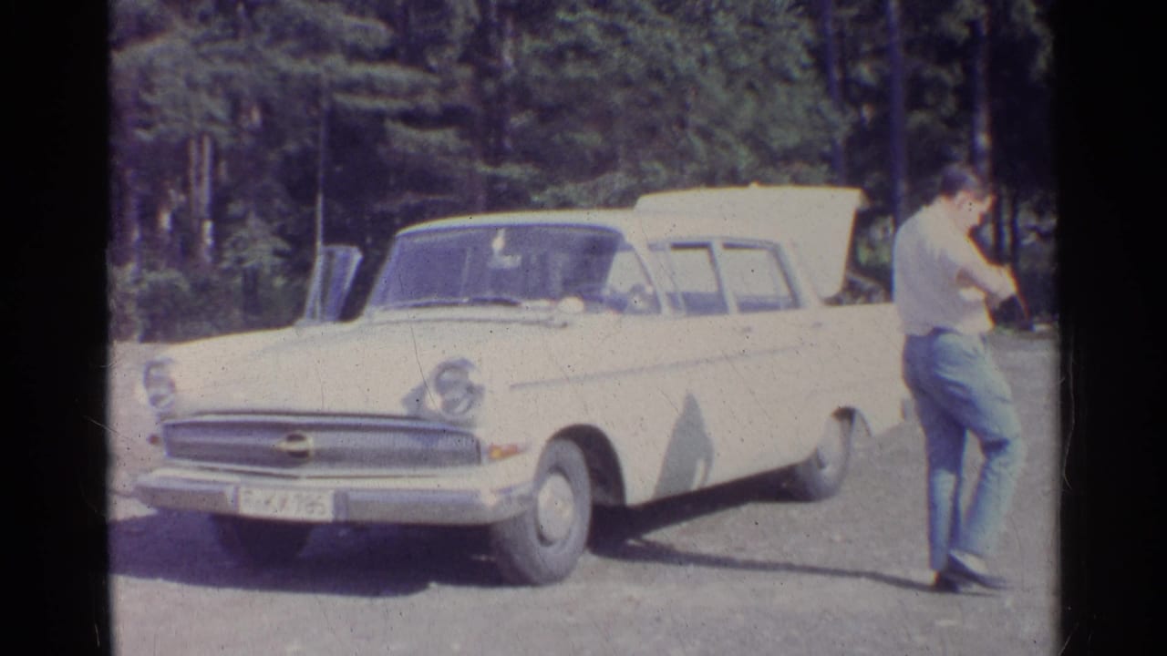 A white vintage sedan sits at a forest pull-off as a man walks past the front of the car in 1961 Germany home-movie footage.