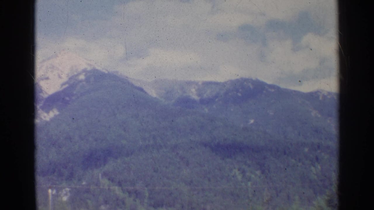 1961 German home-movie still of a snow-flecked mountain range and forested lower slopes under overcast sky.