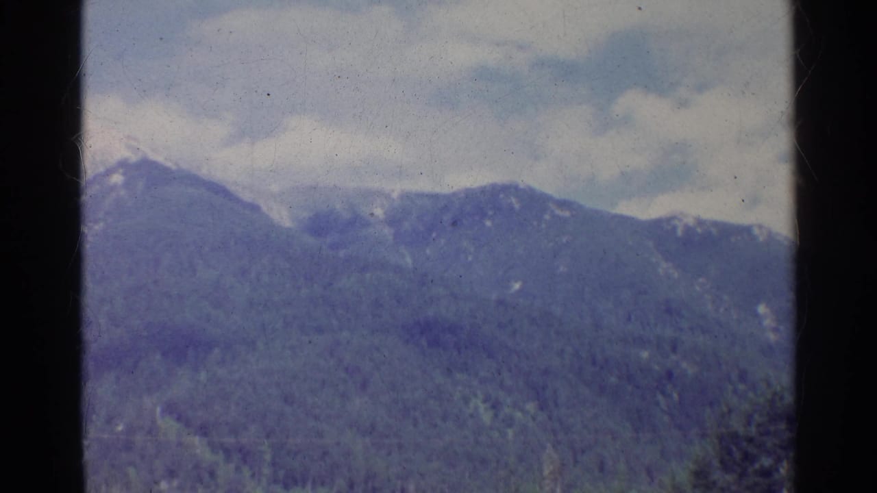 1961 German 8mm still of a forested mountain range with small snow patches and soft cloud cover.