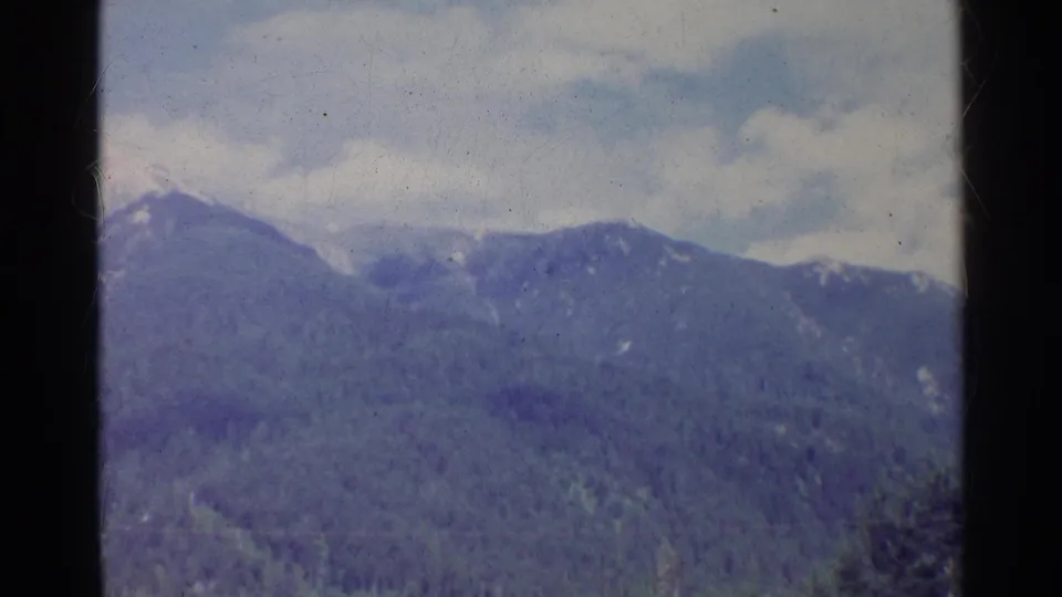 1961 German 8mm still of a forested mountain range with small snow patches and soft cloud cover.
