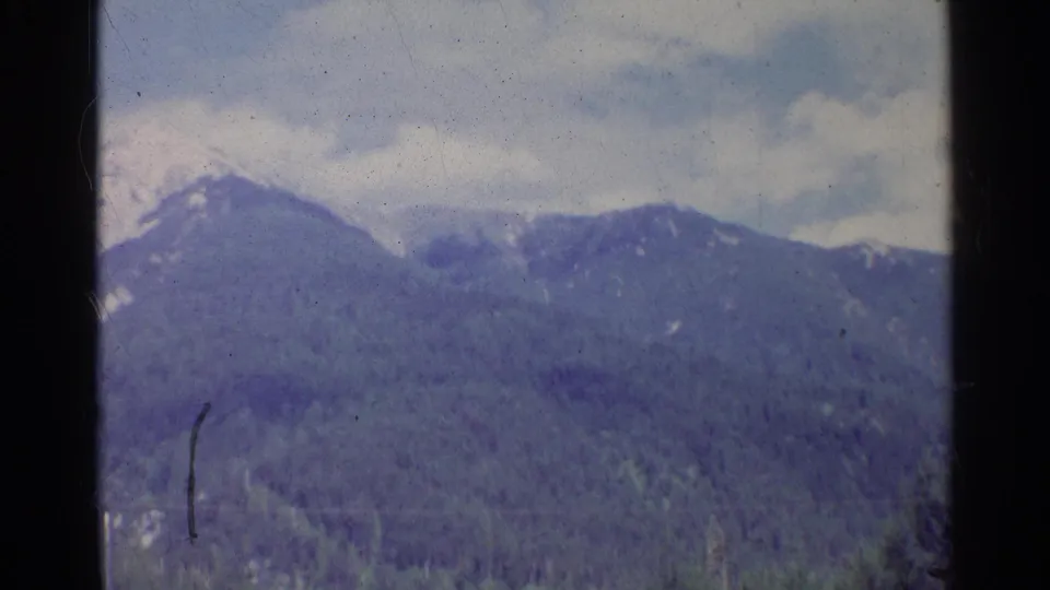 1961 Germany home-movie frame showing distant snowy mountain peaks above forested slopes and a partly cloudy sky.