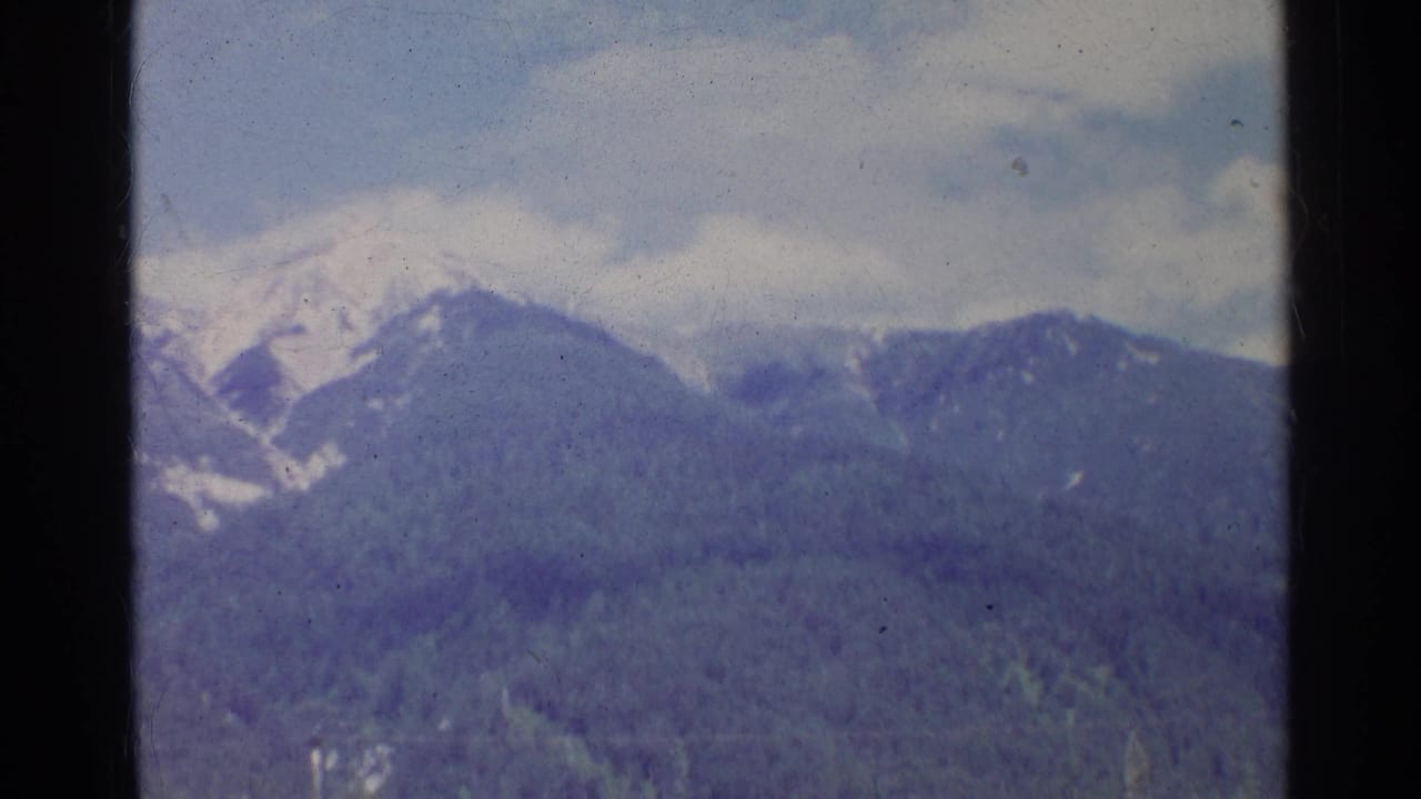 1961 Germany 8mm frame with a snow-bright mountain ridge, dark wooded lower slopes, and soft cloudy sky.