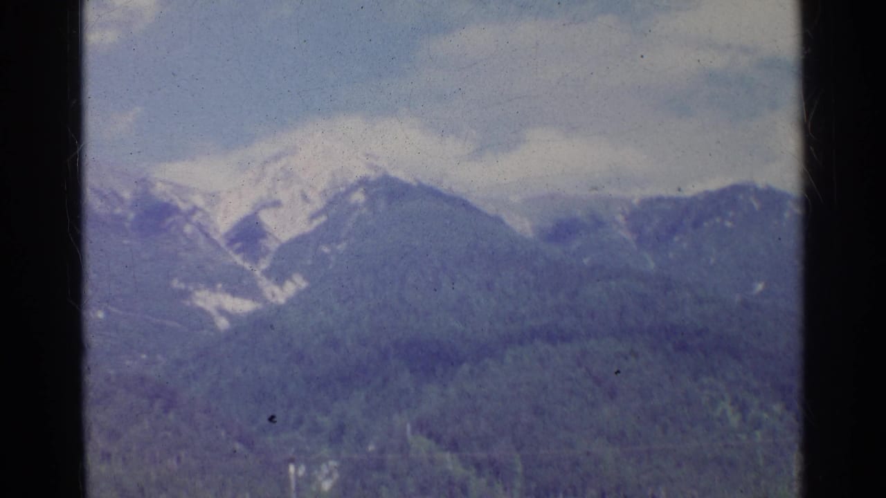 1961 Germany home-movie still showing a snowy peak and blue-toned forested mountain slopes beneath clouds.