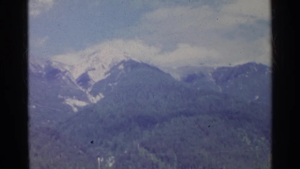 1961 Germany home-movie still showing a snowy peak and blue-toned forested mountain slopes beneath clouds.