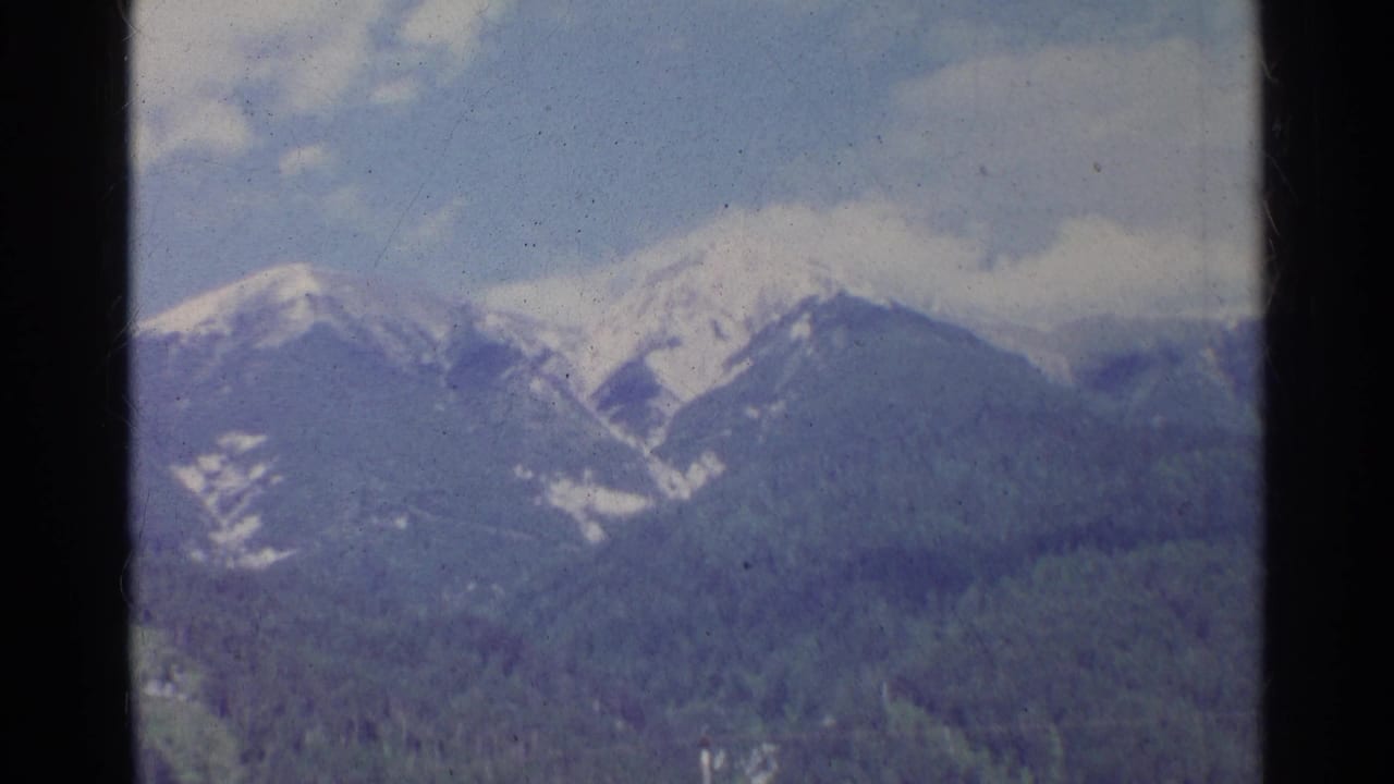 1961 Germany 8mm frame showing snow-capped mountains and forested slopes beneath a blue sky, viewed through a window frame.