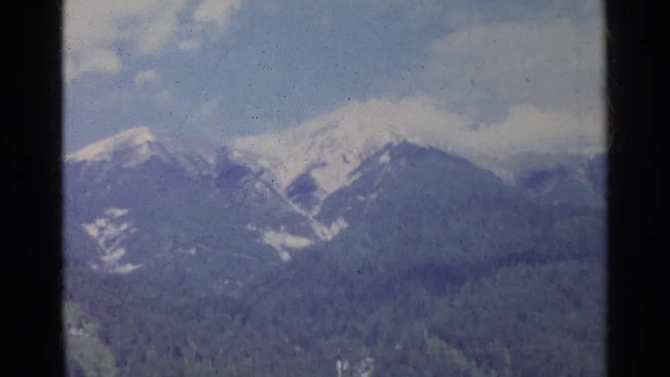 1961 Germany 8mm frame showing snow-capped mountains and forested slopes beneath a blue sky, viewed through a window frame.