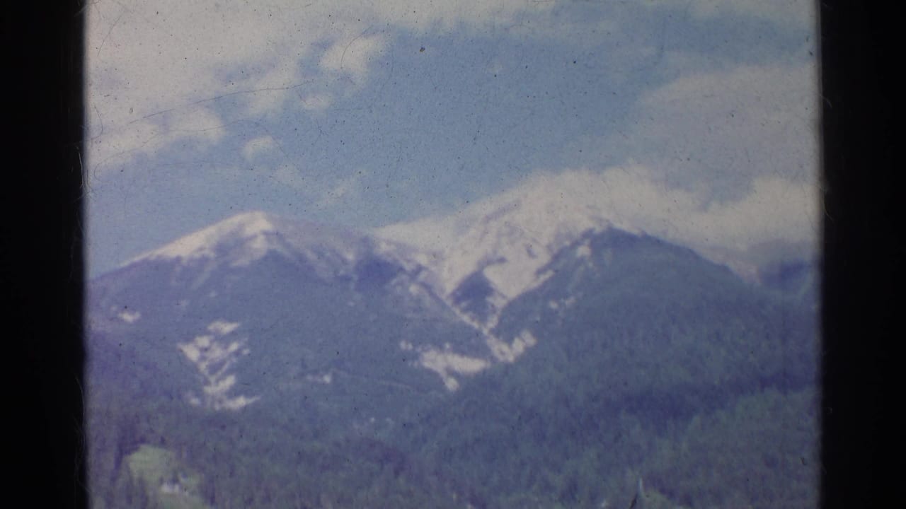 1961 Germany 8mm frame of an alpine mountain range with snow on the peaks and dense forest below.