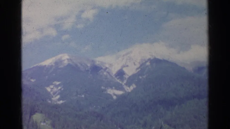 1961 Germany 8mm frame of an alpine mountain range with snow on the peaks and dense forest below.