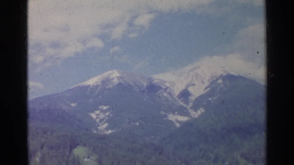 1961 Germany 8mm frame showing snow-covered peaks behind forested foothills, with soft color and visible film wear.