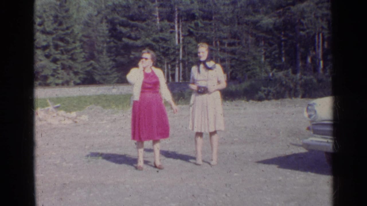 Two women in 1960s-style dresses stand outdoors in a German mountain setting, with forest behind them and a car partly visible.