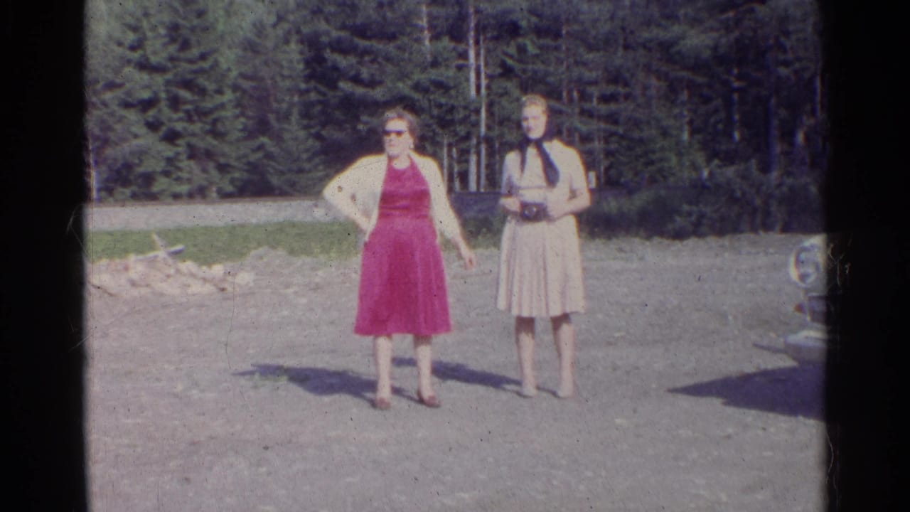 1961 Germany home-movie frame of two women in period dresses standing in bright sun near a forested roadside or clearing.
