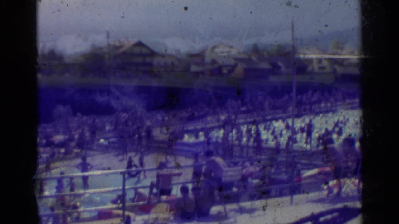 Busy outdoor swimming pool scene in 1961 Germany, filmed from above with bathers, pool railings, trees, and distant structures.