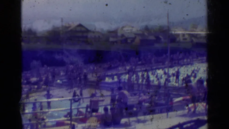 Busy outdoor swimming pool scene in 1961 Germany, filmed from above with bathers, pool railings, trees, and distant structures.