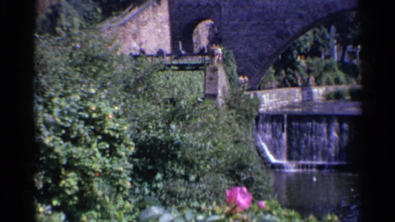 1961 Germany frame showing the bridge arch, garden plantings, a small dam, a stream, and a tiny distant person near a railing.