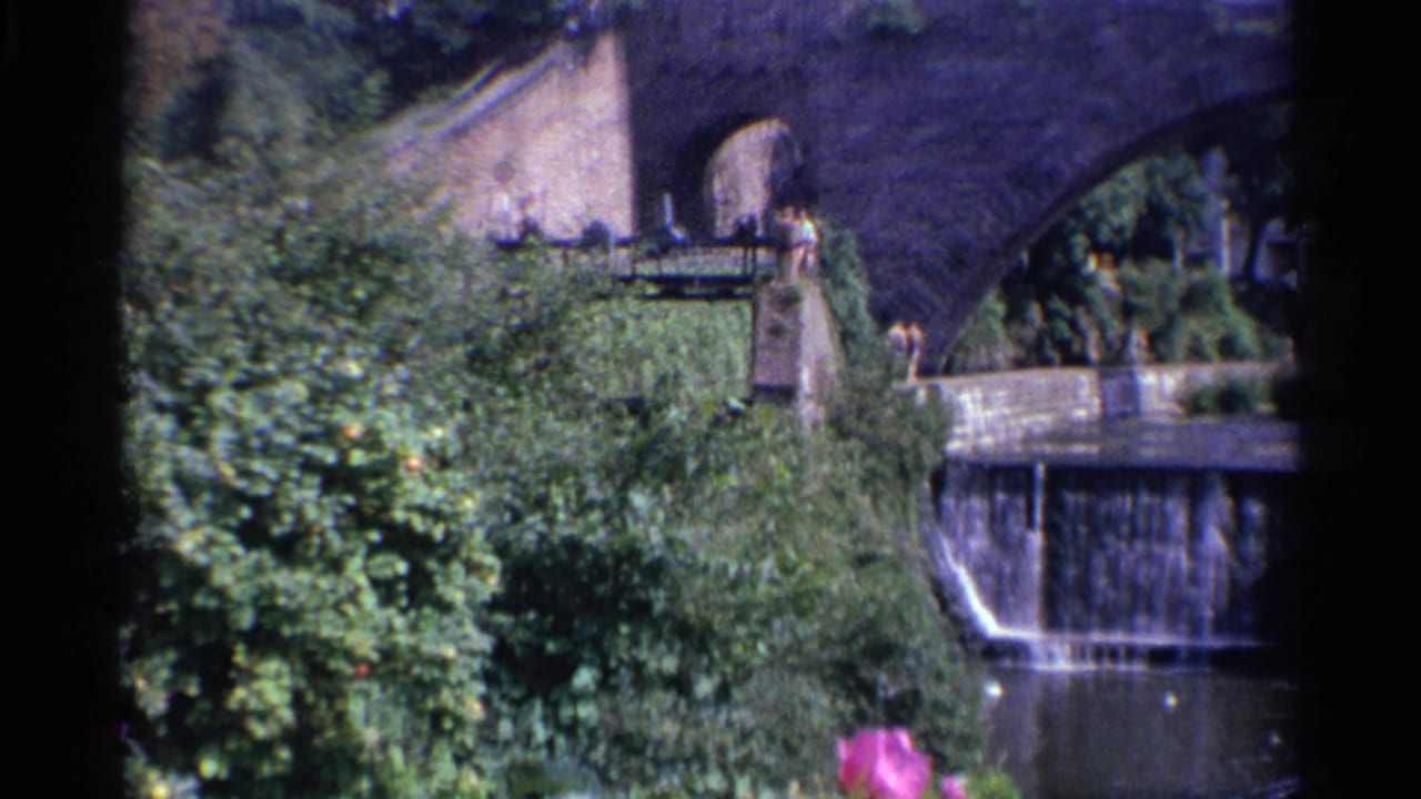 1961 Germany 8mm frame showing a stone arched bridge, lush greenery, a yellow building above, and water spilling into a river.