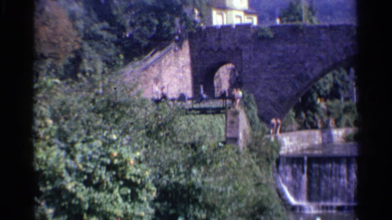 1961 Germany 8mm frame with a stone bridge arch, dense ivy and trees, a hillside building, and a low waterfall below.