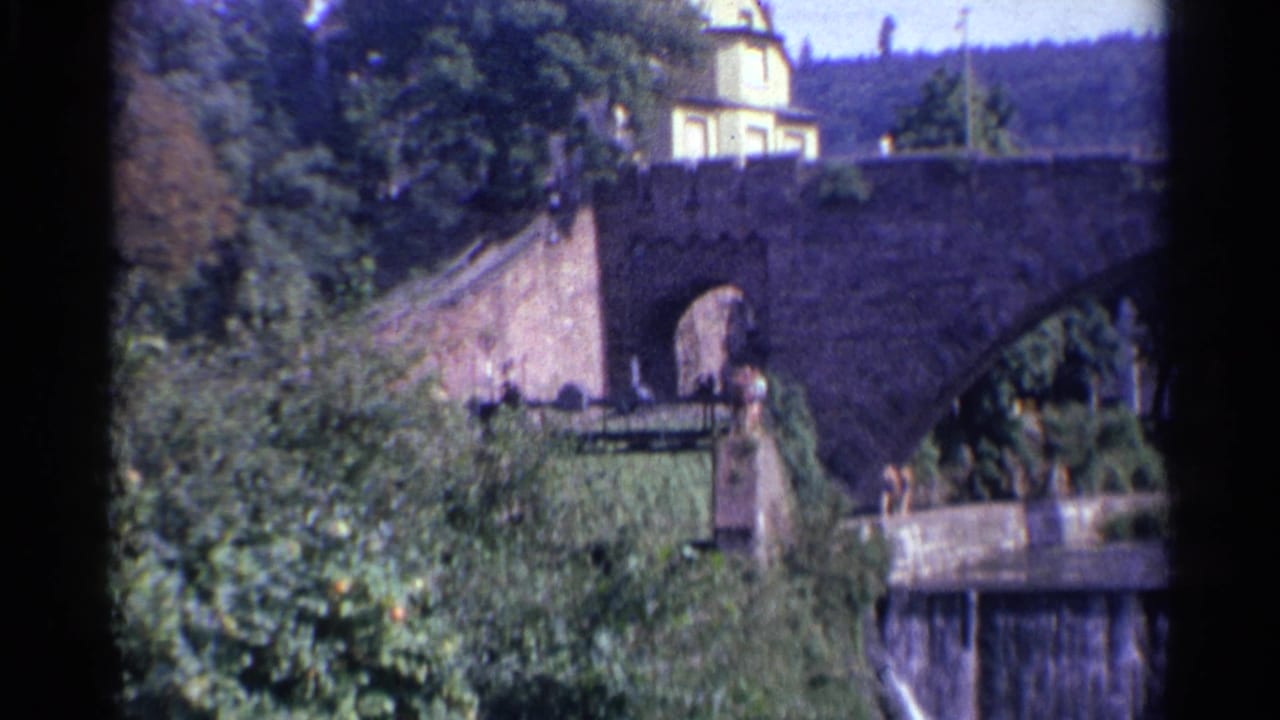 1961 Germany 8mm frame showing the upper span of a stone bridge, a yellow building beyond it, ivy, trees, and water below.