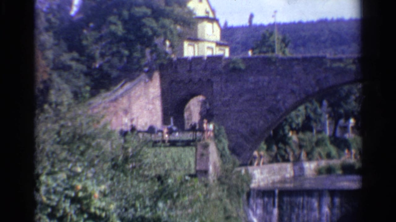 1961 Germany 8mm frame showing a sunlit stone bridge, ivy-covered bank, yellow building, and small waterfall in a wooded river scene.
