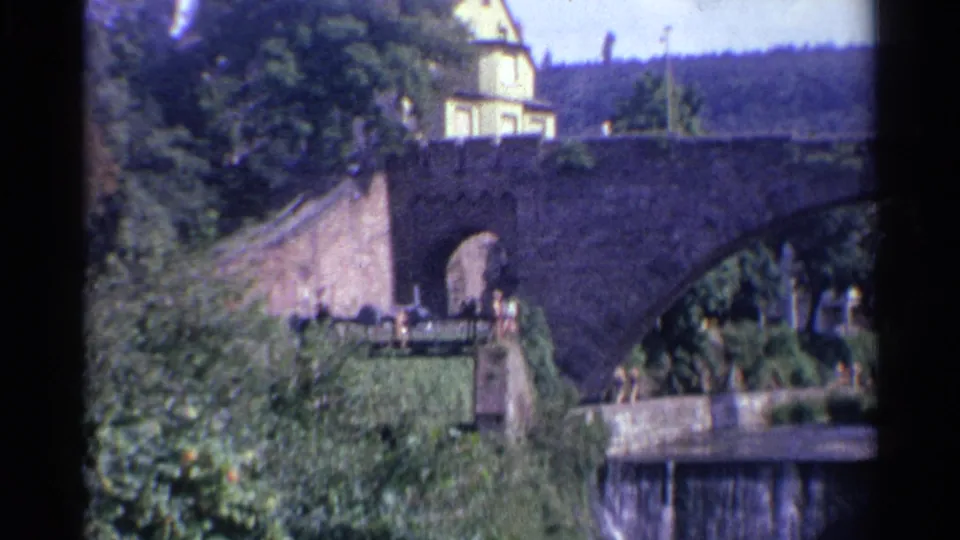 1961 Germany 8mm frame showing a sunlit stone bridge, ivy-covered bank, yellow building, and small waterfall in a wooded river scene.