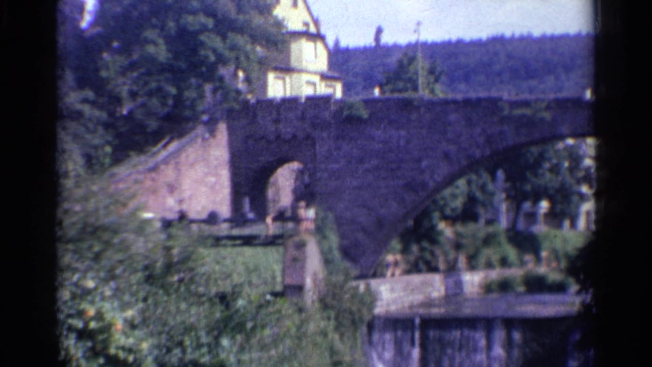 1961 Germany home-movie frame showing a stone arch bridge, greenery, a riverside walkway, distant people, and a pale building beyond.