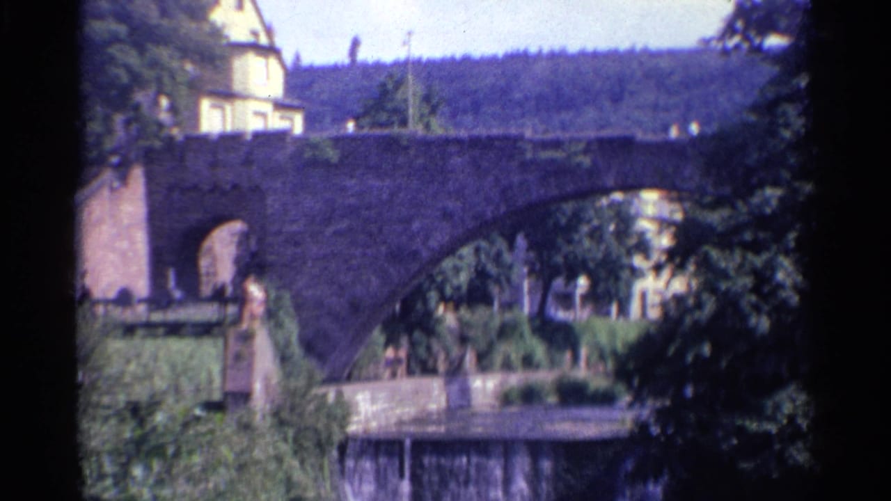 1961 Germany footage frames the large stone bridge arch with dense trees, trimmed greenery, a river edge, and distant people near the bank.