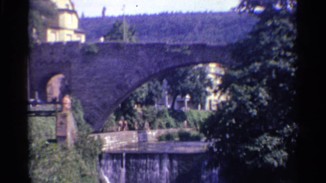 Final 1961 Germany frame shows the stone bridge, shaded trees, riverfront landscaping, and a pale building partly visible behind the parapet.