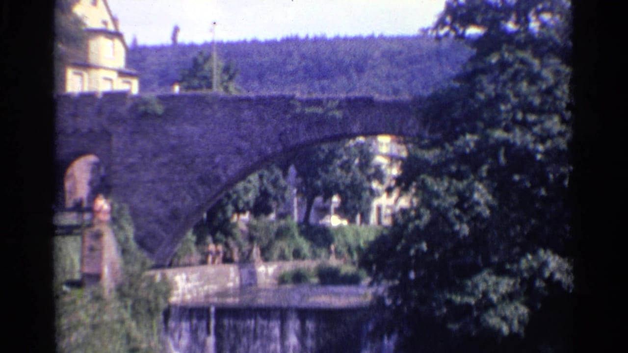 Stone arch bridge with trees, hillside building, and small pedestrians near the riverside.