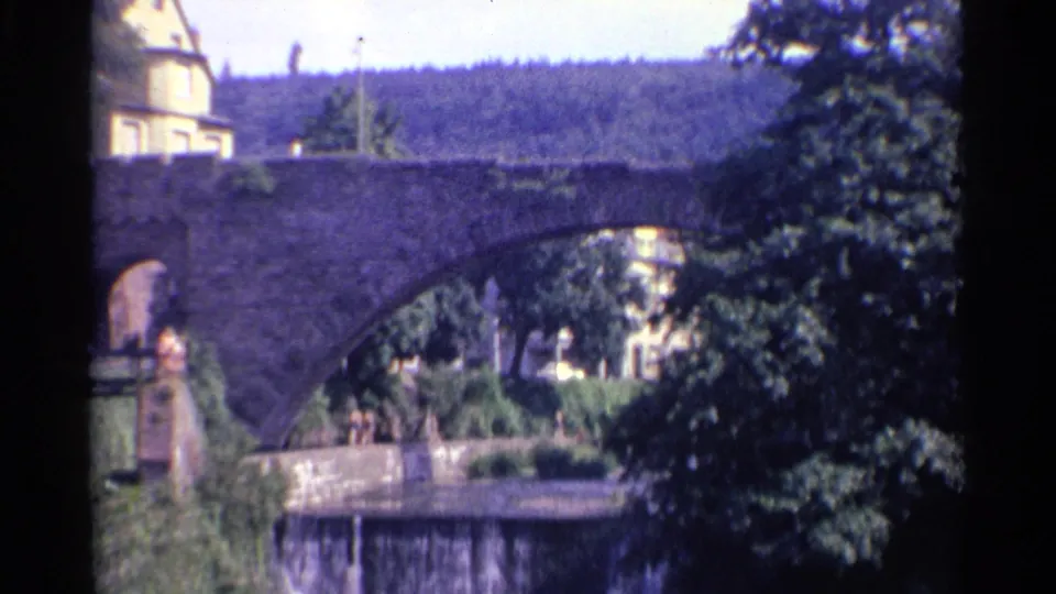 1961 Germany 8mm frame showing a stone arch bridge framed by trees, with a pale building above and distant pedestrians near the water.