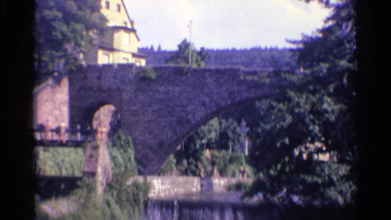 A soft 8mm view of a German stone bridge in 1961, with pedestrians at the bridge approach and dense trees partly covering the frame.