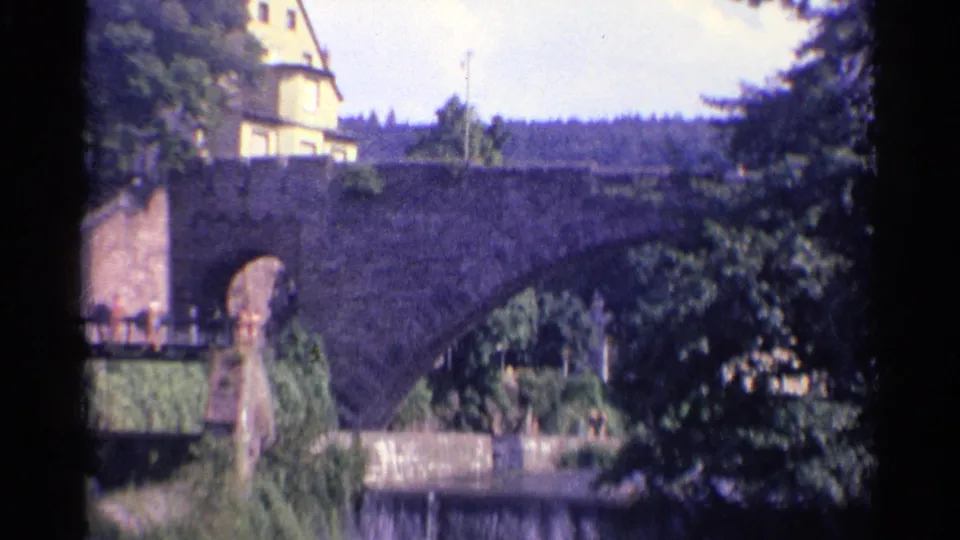 A soft 8mm view of a German stone bridge in 1961, with pedestrians at the bridge approach and dense trees partly covering the frame.
