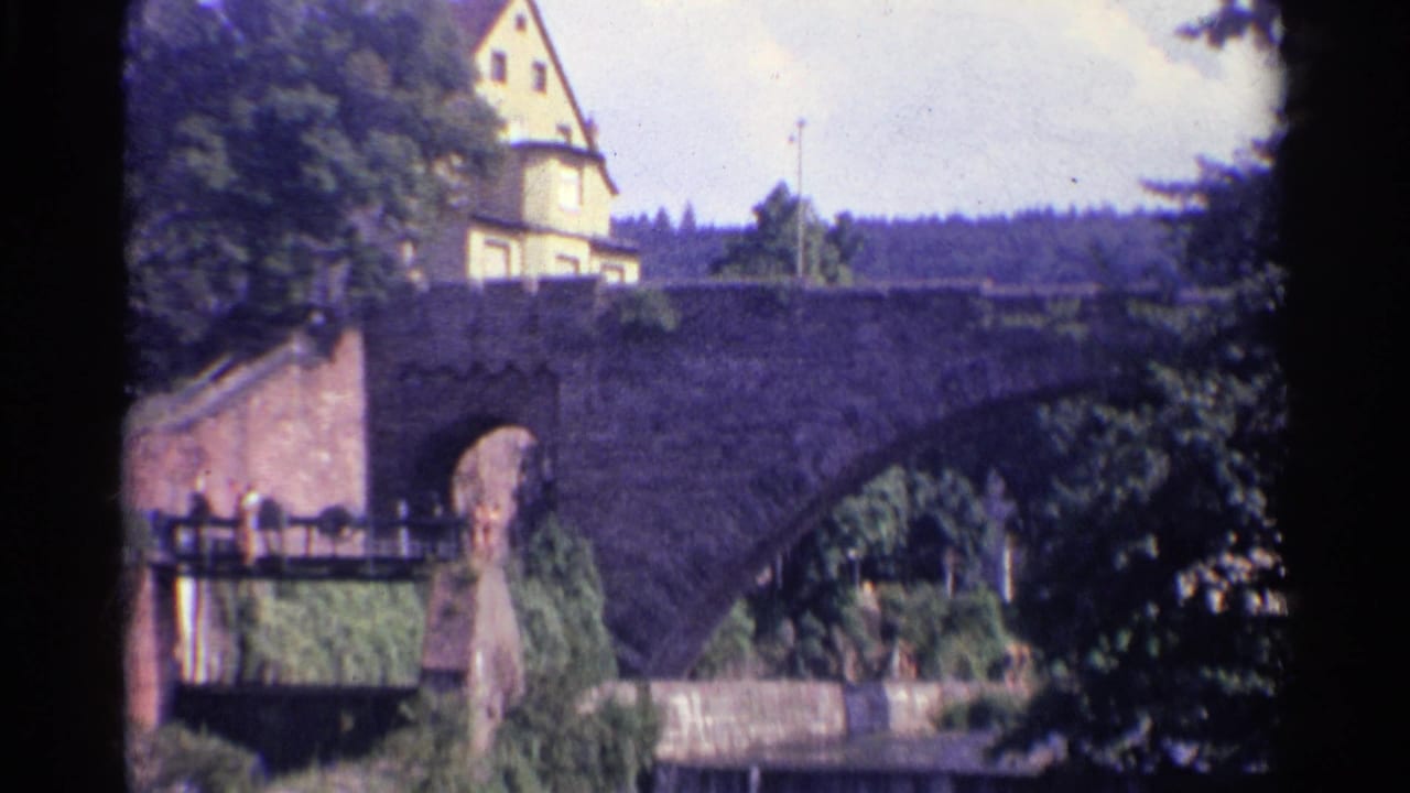 1961 Germany home-movie frame showing the bridge approach, masonry arch, leafy trees, a hillside building, and small pedestrians on a walkway.