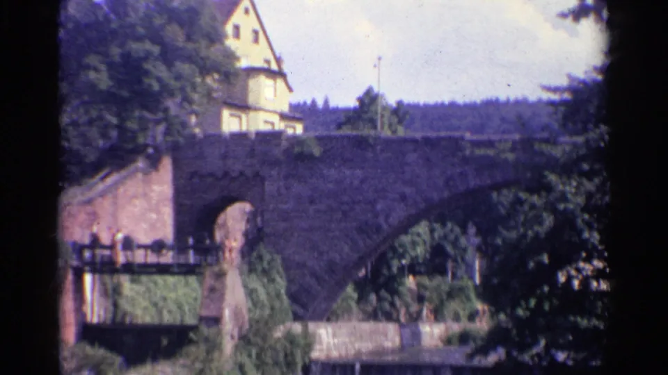 1961 Germany home-movie frame showing the bridge approach, masonry arch, leafy trees, a hillside building, and small pedestrians on a walkway.