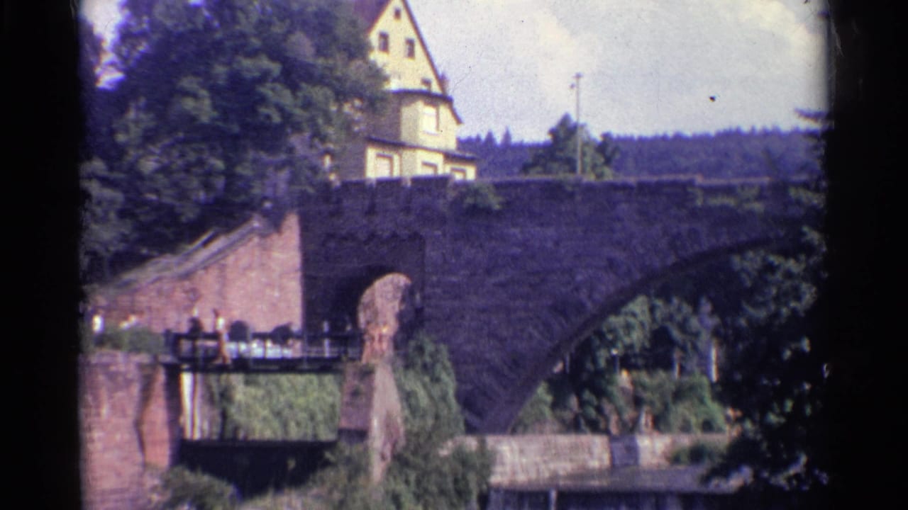 Final 8mm frame of the German bridge view, with the large stone arch, riverside vegetation, hillside building, and small pedestrians at left.