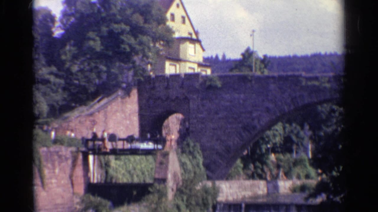 Stone arch bridge with lower walkway, trees, and a yellow building above.