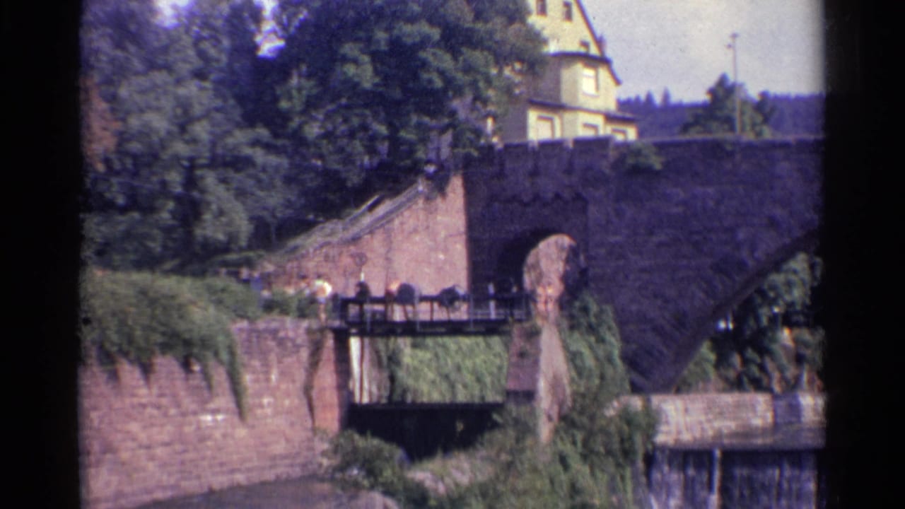 1961 Germany home-movie frame with a pedestrian on a lower walkway, overgrown stone walls, trees, and the main arch bridge above.