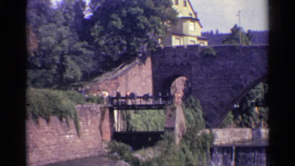 1961 Germany home-movie frame with a pedestrian on a lower walkway, overgrown stone walls, trees, and the main arch bridge above.