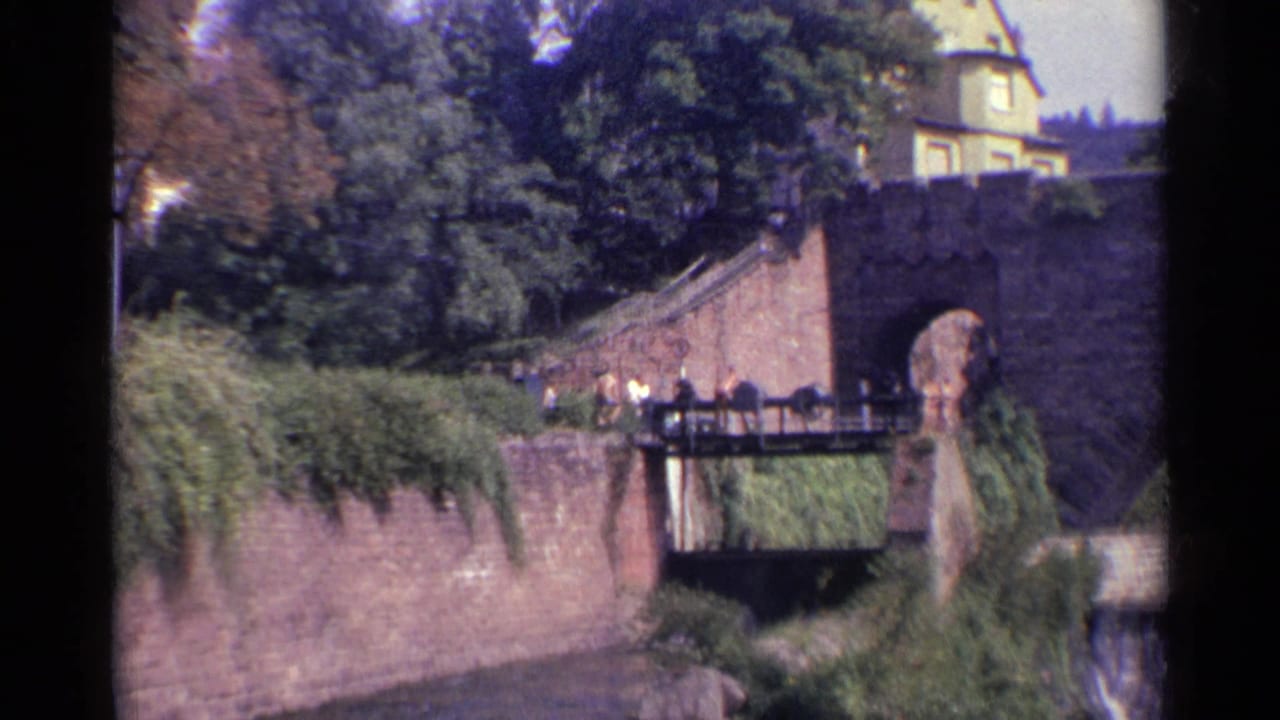1961 Germany home-movie frame showing a stone riverside wall, small footbridge, pedestrians, dense trees, and a building above the riverbank.