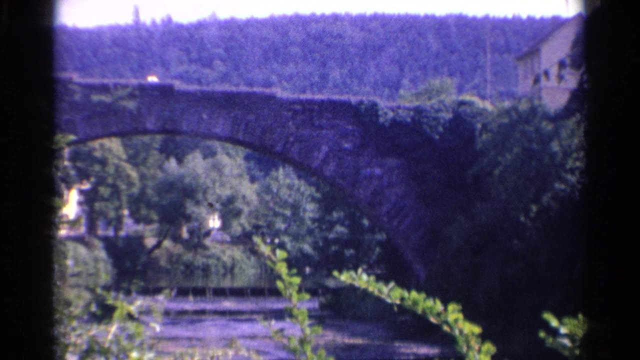 1961 Germany home-movie view of a dark stone arch bridge over the river, with dense greenery, a hillside, and a riverside platform below.