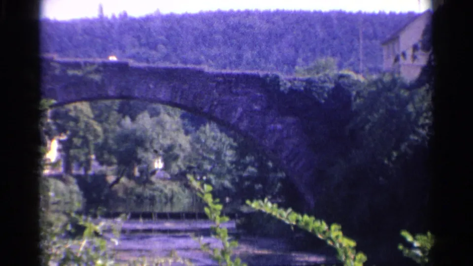 1961 Germany home-movie view of a dark stone arch bridge over the river, with dense greenery, a hillside, and a riverside platform below.