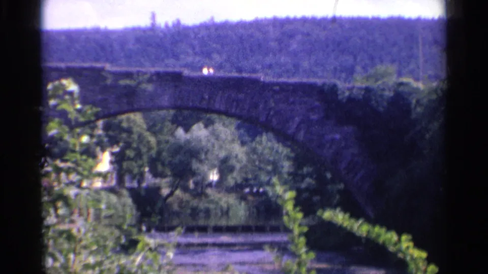 1961 Germany 8mm frame showing a stone arch bridge over a river, partly obscured by leaves, with a forested hillside in the distance.