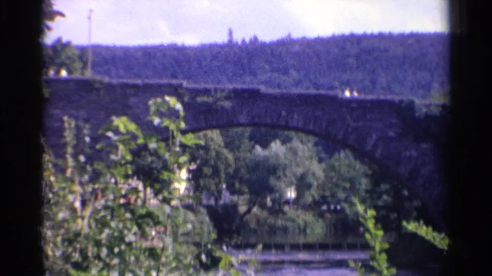 1961 Germany home-movie view of a stone bridge crossing a river, framed by leaves and backed by a tree-covered hill.