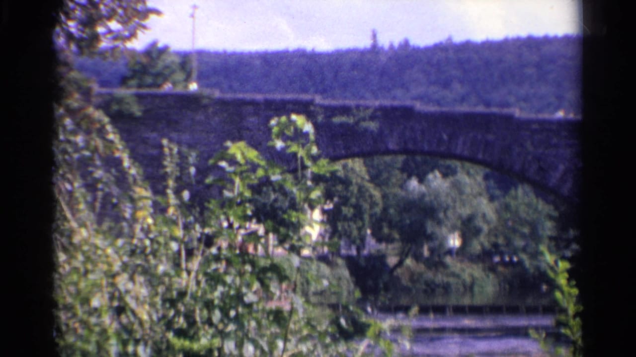 1961 Germany 8mm frame with the stone arch bridge centered behind green foliage, river water below, and wooded hillside beyond.