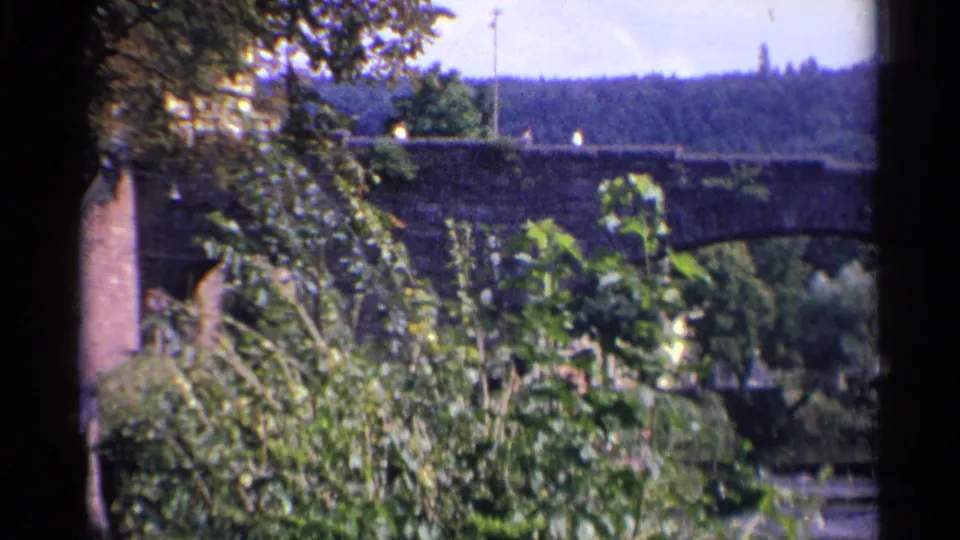 1961 German 8mm frame showing bushes in the foreground and a stone bridge crossing a river or stream beyond.