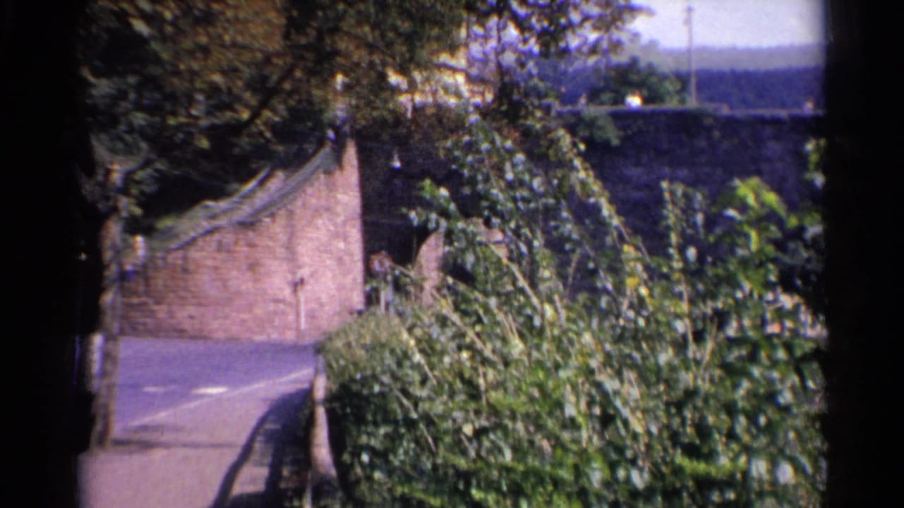 1961 Germany travel footage frame with a curving paved road, masonry wall, bushes, and a bridge beyond.