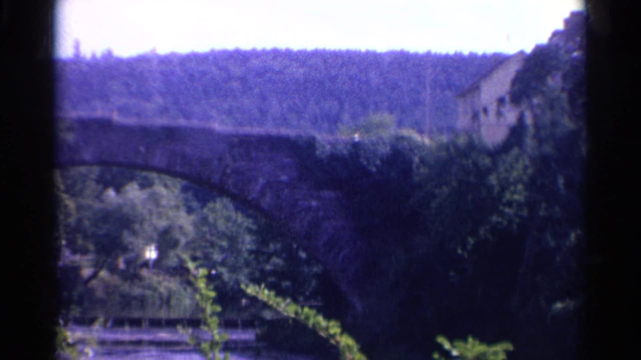 1961 8mm view of a stone arch bridge below a wooded hillside, with a building partly visible at the right edge.