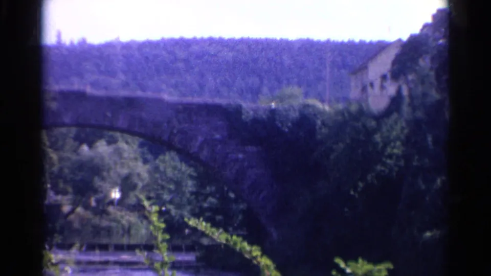 1961 8mm view of a stone arch bridge below a wooded hillside, with a building partly visible at the right edge.
