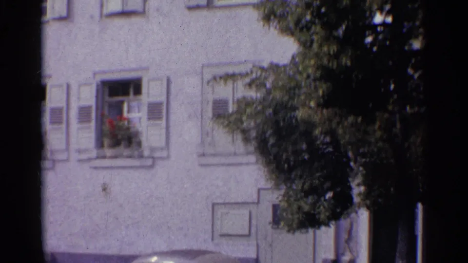 1961 Germany 8mm frame of a white stucco building facade with pale shutters, flower boxes, and tree branches along the right edge.