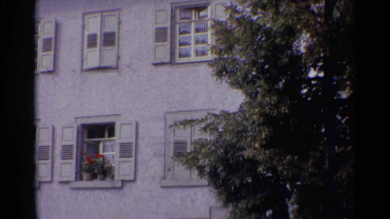 1961 Germany 8mm frame showing shuttered windows on a white stucco building, with a red flower box and dark tree foliage in the foreground.