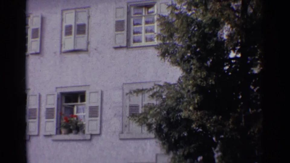 1961 Germany 8mm frame showing shuttered windows on a white stucco building, with a red flower box and dark tree foliage in the foreground.